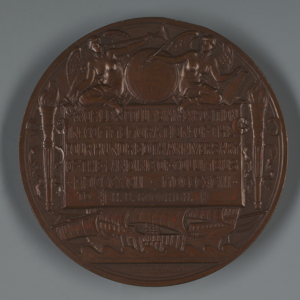 A large circular bronze medal with shallow relief designs, including two winged angels at top, sailing ships below, and a central text panel about the World’s Columbian Exposition.