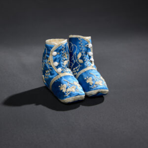 A pair of delicate blue silk children’s booties with white edging and white leaf appliques and detailing. Each bootie has three white buttons on the front of the ankle.