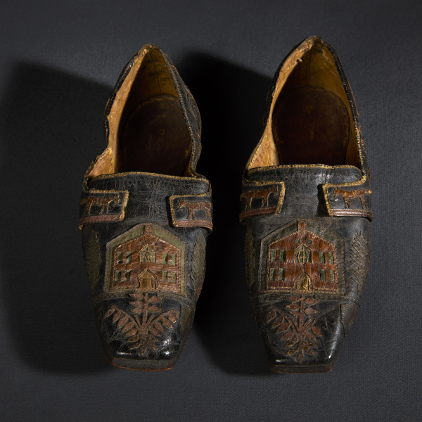 A pair of black leather shoes decorated with detailed stitched leather appliques in the shape of houses, plants, and animals. The interior of the shoe is natural brown leather.