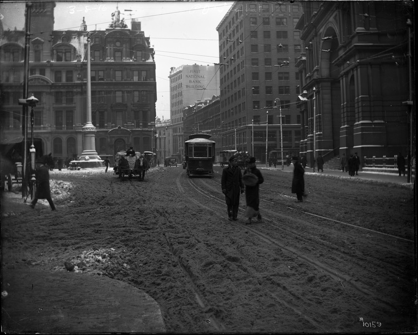 Street scene (winter), snow on the ground. Horse drawn wagon, trolley #91, sign: South Station, North Station, First National Bank Building, sign: left: New England Mutual Life Insurance Building, Angel Memorial Park, Far Right: Post Office + Subtreasury. Building, pedestrians.