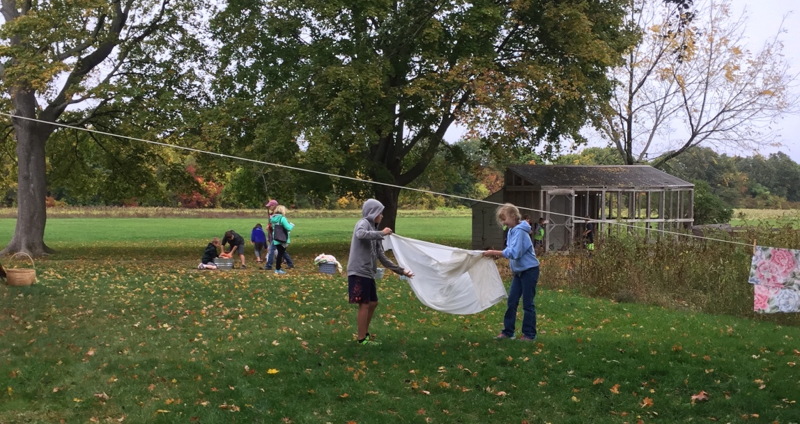 Doing the laundry at Stekionis House In the foreground, two students prepare to hang a wet sheet on a laundry line. In the background, students work with washboards over buckets, doing the laundry