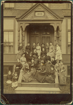 A group of young women on the stairs leading to the front entranceway of a building with a plaque that say 1883. Many of them hold tennis racquets.
