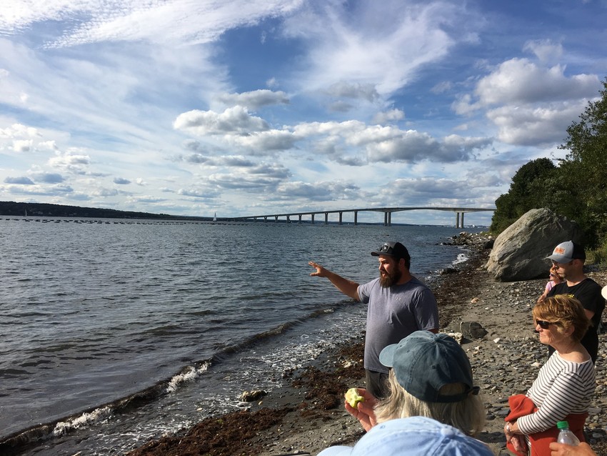 Man on beach giving a tour. Bay in foreground, bridge in distance.