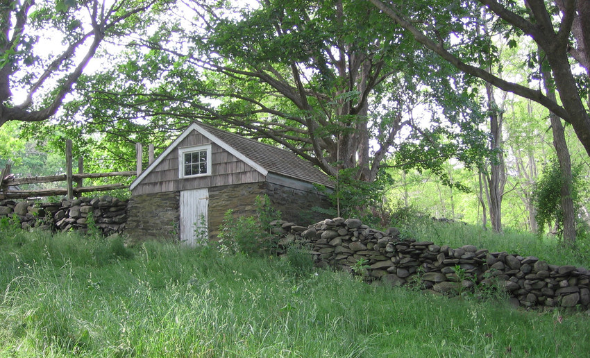 Small stone and shingled outbuilding in a field flanked by stone wall.