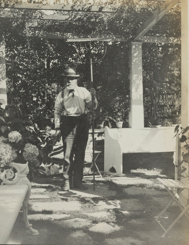 Black and white photograph of a man with a white beard, wearing and hat and holding a rake.