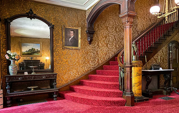 Red carpeted stairway with ornate wallpaper and woodwork.