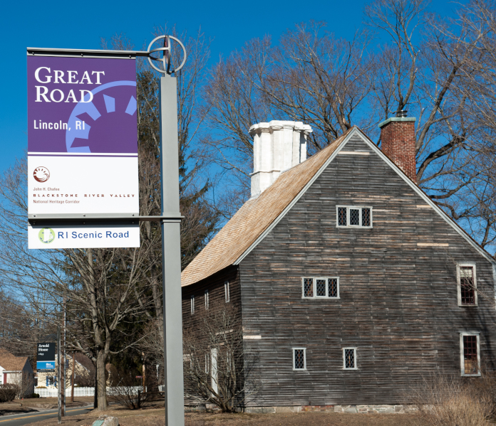 Historic wooden house with a steep roof beside a Great Road, Lincoln, RI sign, bare trees surround under a clear sky.