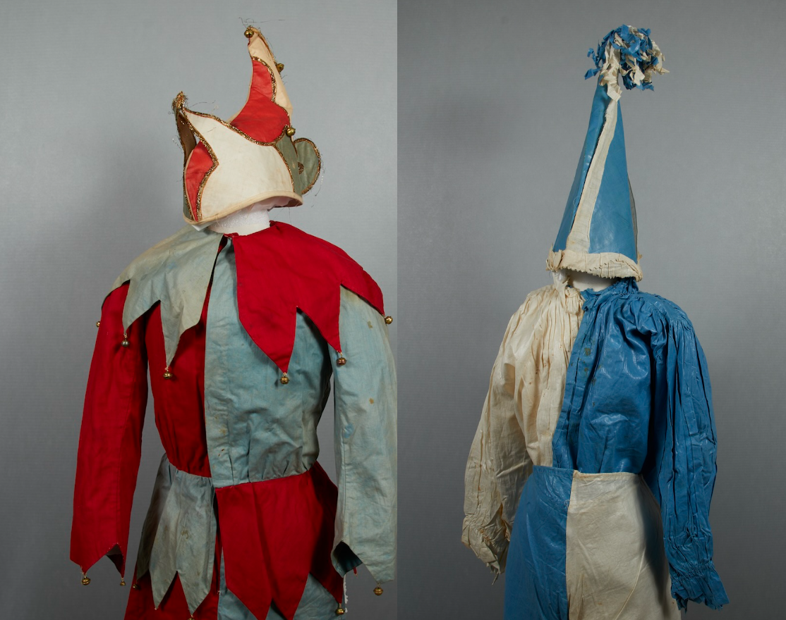 The images shows two costumes. On the left is a jester costume fabricated from red and pale blue fabric and a pointed, multi-colored hat with bells. On the right is a blue and white clown suit with a tall, pointed hat.
