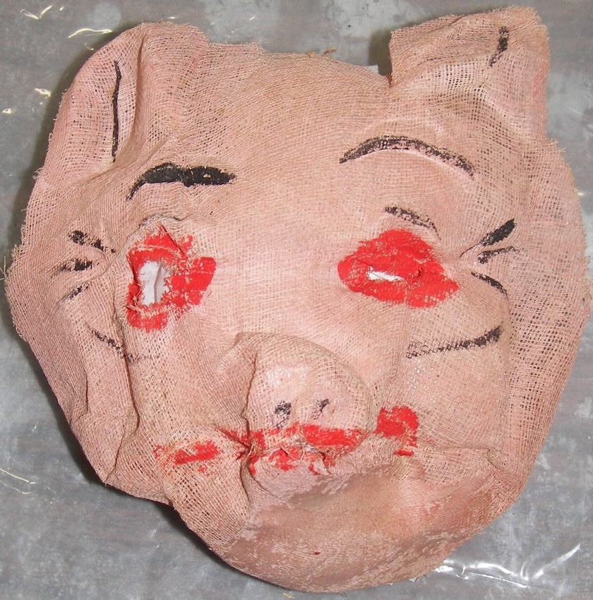 A worn and crumpled pig mask made of rough fabric. The mask features red lips, black eyebrows, and a pig-like nose. The material is frayed and tattered around the edges, with holes for the eyes and some of the painted areas smudged or chipped.