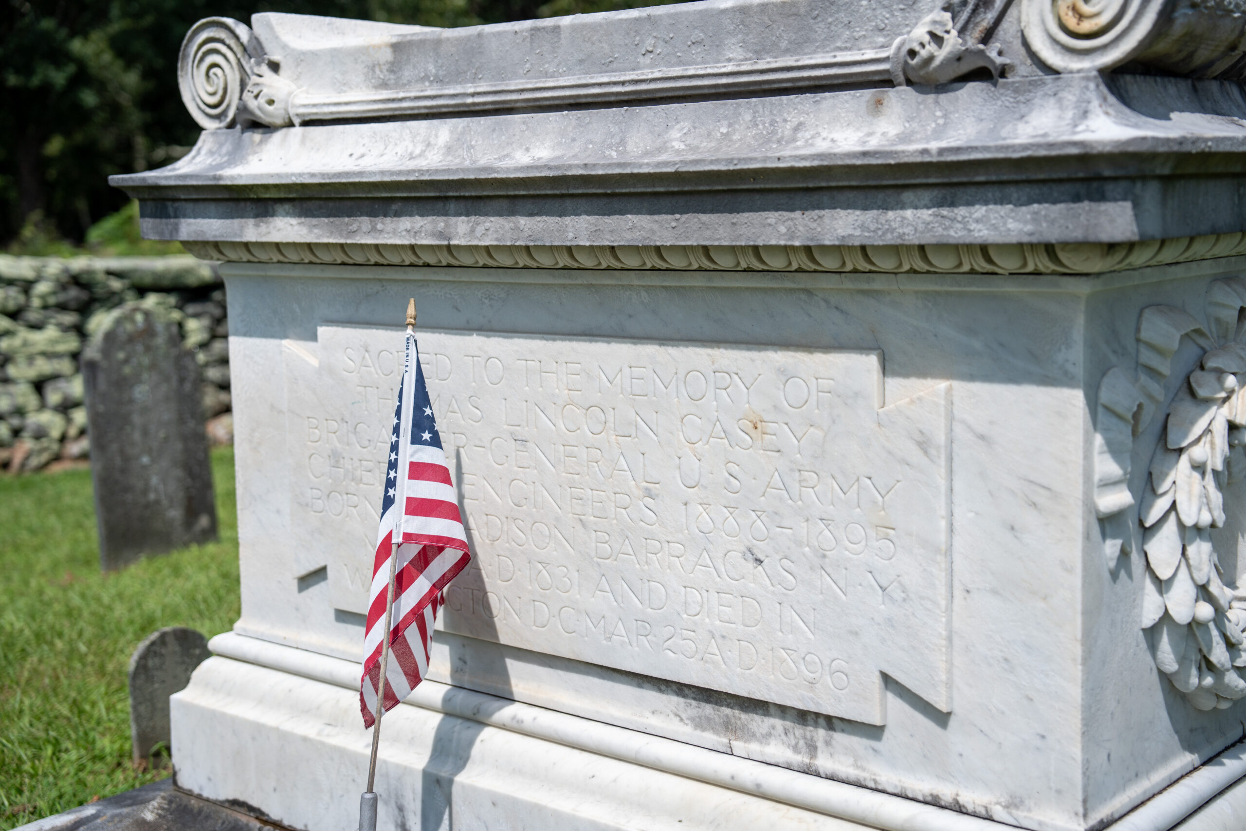 Ornate gravestone for Thomas Lincoln Casey features laurels and oak wreaths. Epitaph is partially obscured by an American flag.