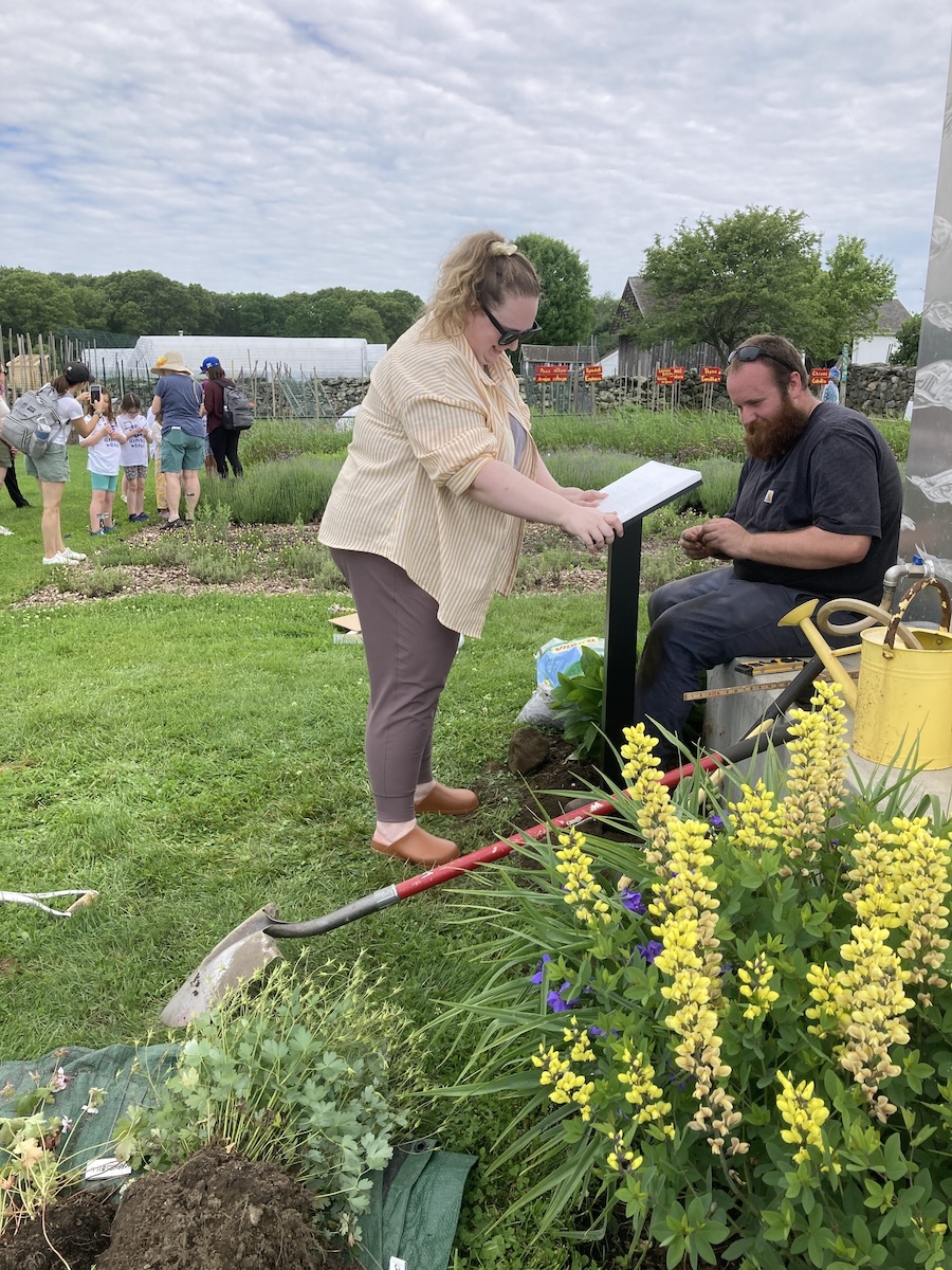 Outdoor scene at a farm. In the foregorund, a person is standing and installing a sign. Another is sitting and watching.