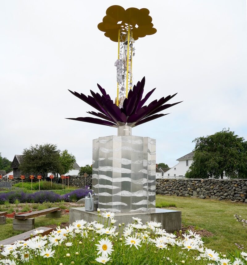 Tall sculpture in an outdoor setting with a field of daisys in the foreground and a stone wall behind it.