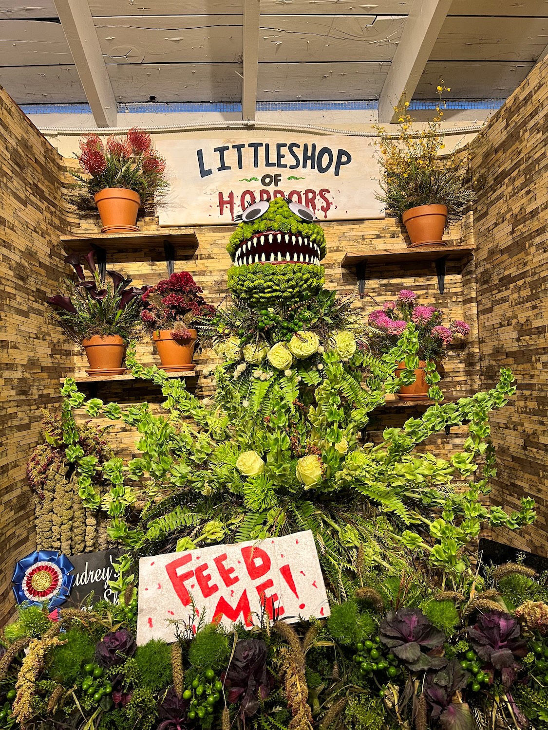 A monster made out of flowers is in small booth with signs that read Feed Me and Little Shop of Horrors.