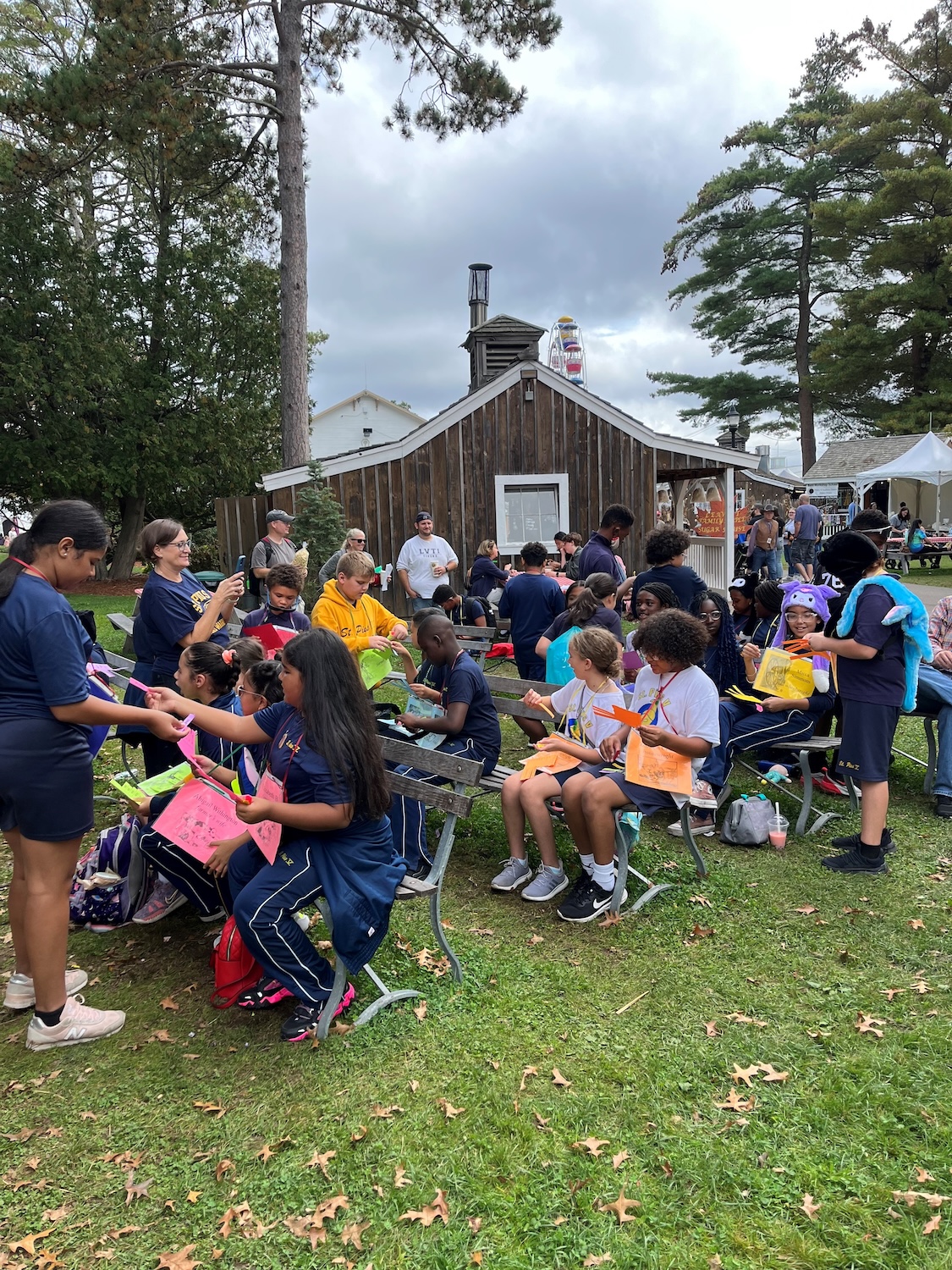 A group of schoolchildren sit on wood benches looking at handouts while adults stand nearby.