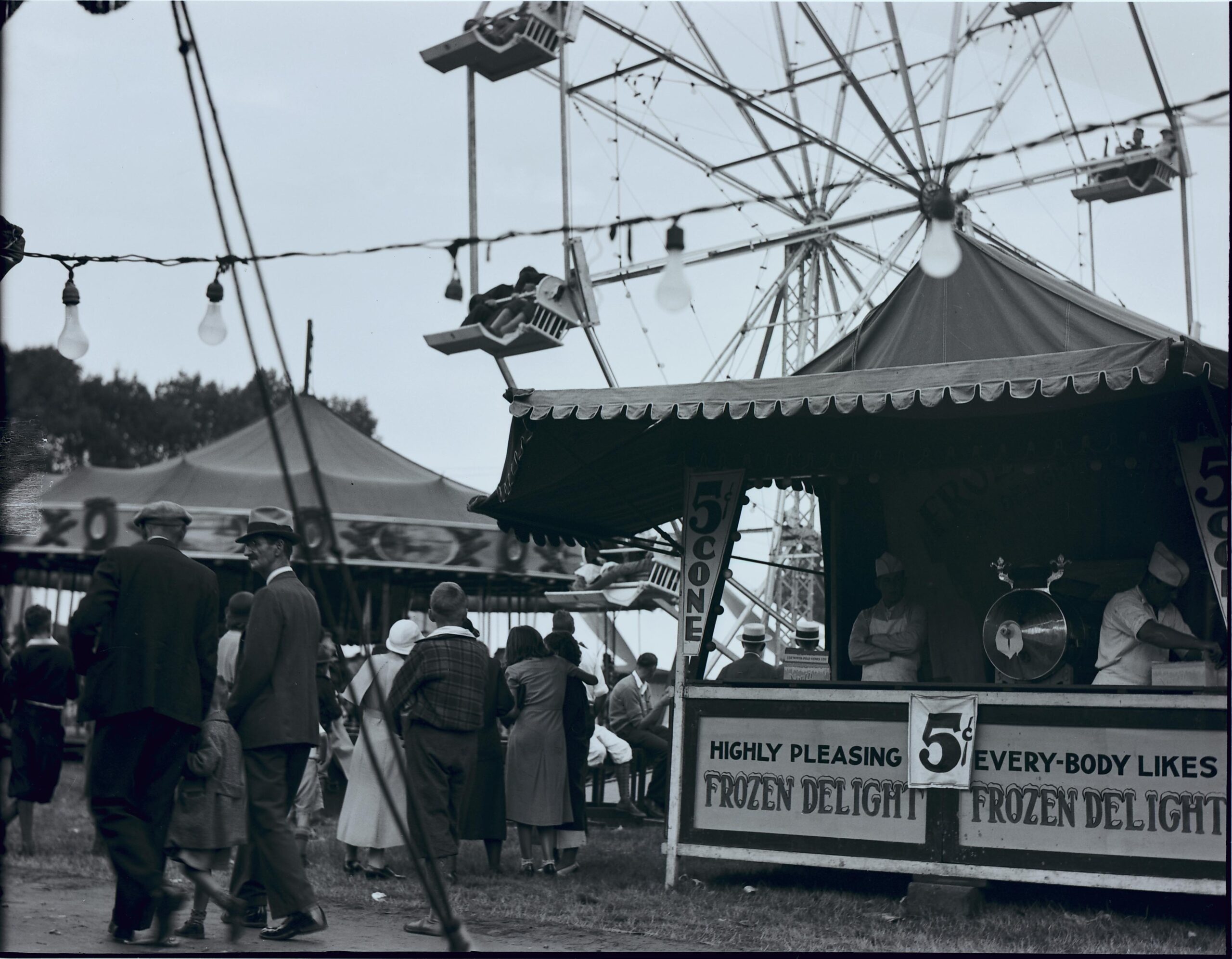 Black-and-white photograph of a fair depicting a ferris wheel, merry-go-round, ice cream stand, and several people walking around.