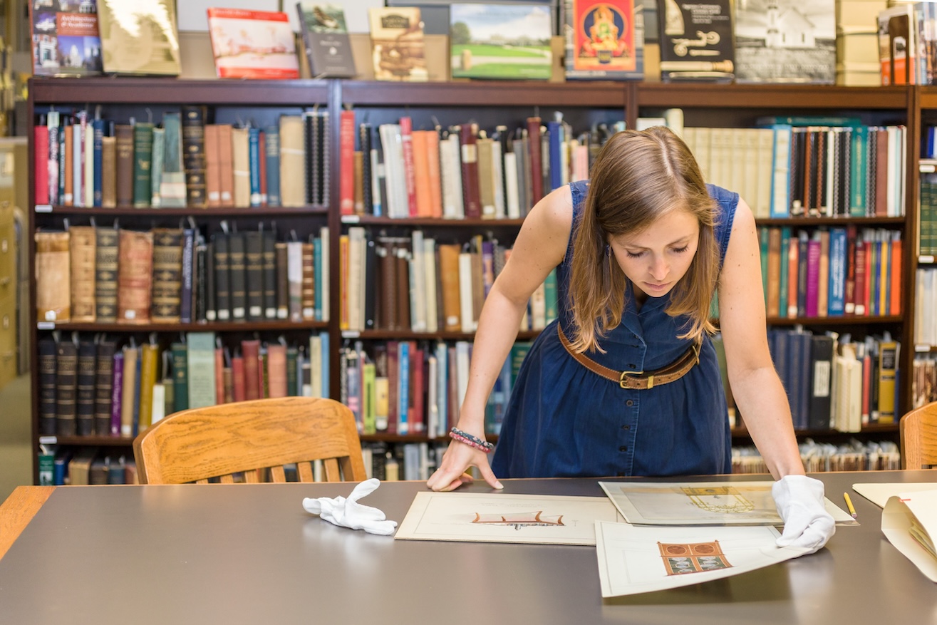 A woman is in a library with books behind her. She leans over a desk, looking closely at sketches.