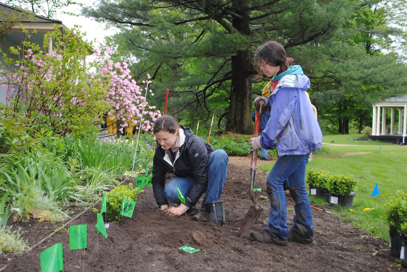 A person kneels in the dirt next to a flower bed planting a bulb, while another person stands nearby holding a shovel.