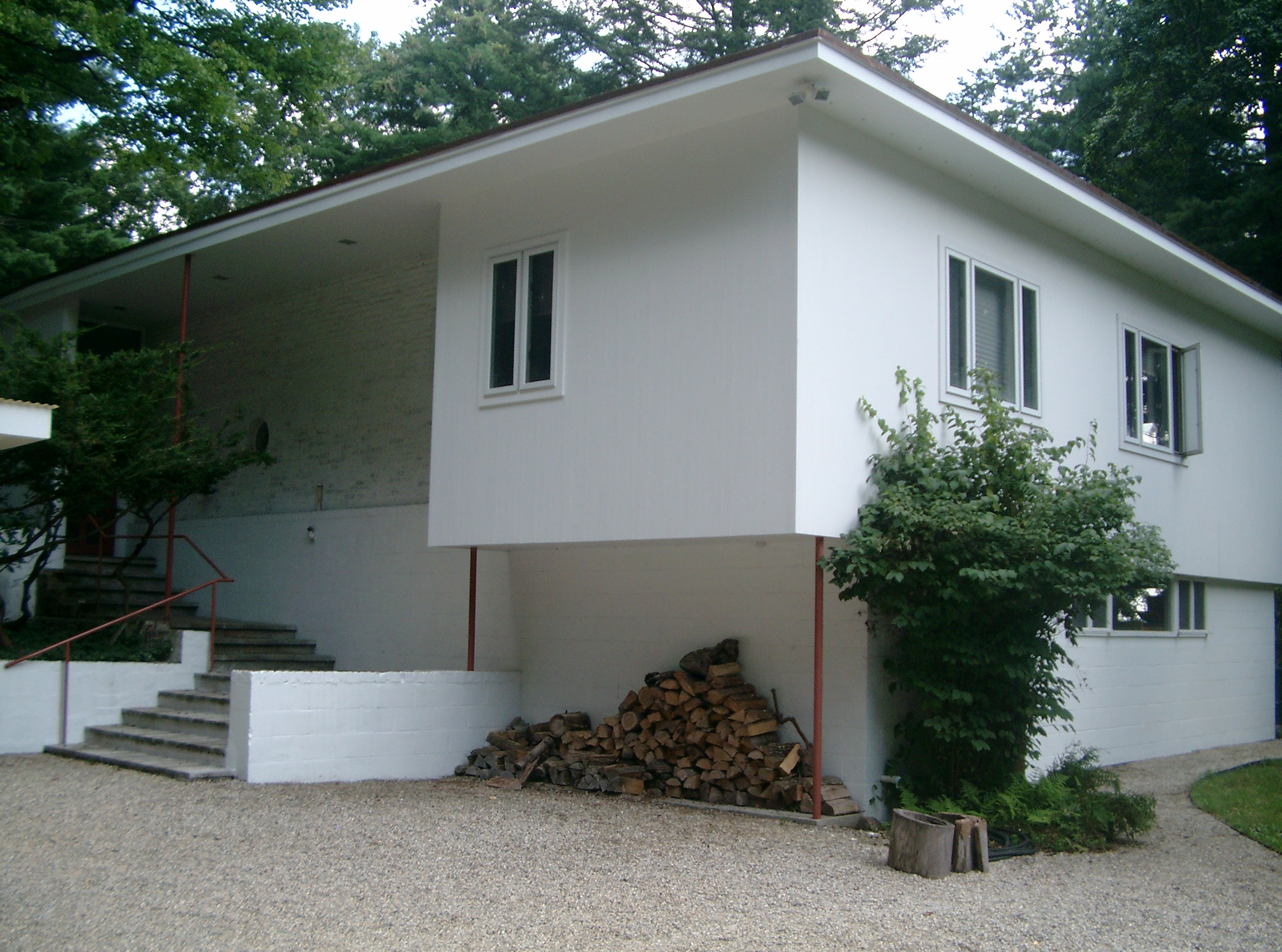 A modern white house with rectangular windows, elevated on stilts, with firewood piled underneath.