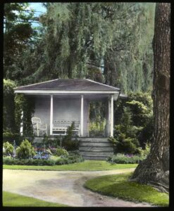 Color photograph of a garden house with rattan seating. A weeping willow is in the foreground and there is an open door leading into a garden.