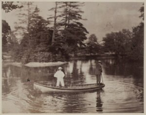 Man and young boy in a boat on a pond. Man is standing at the stern of the boat with a pole in the water to steer the boat. a small island with pine trees is in the distance.