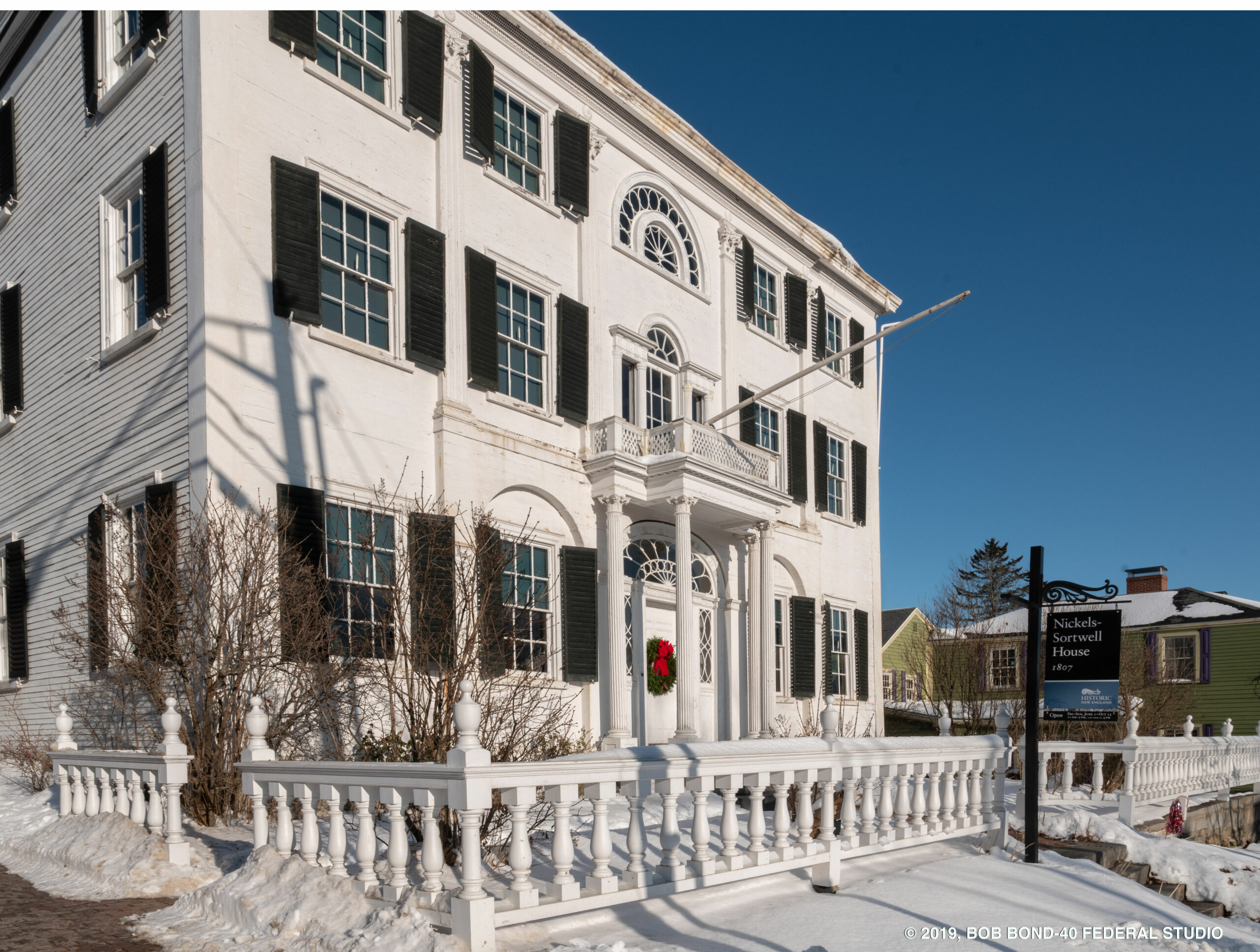 Three-story white house with green shutters and a portico, surrounded by snow and a white fence.