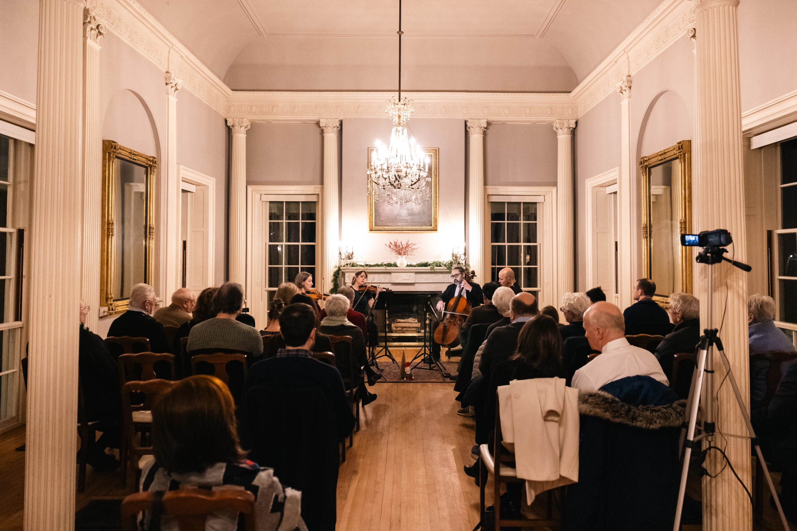 String ensemble performing in a sophisticated room with a chandelier, fireplace, and audience.