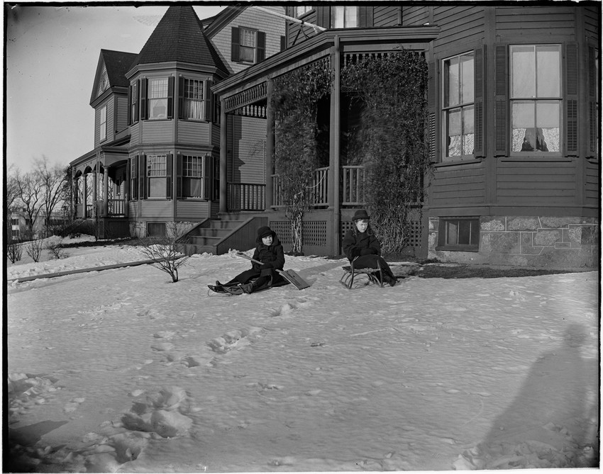 Two children sit on sleds in the snow outside a Victorian-style house with a porch and decorative plants.