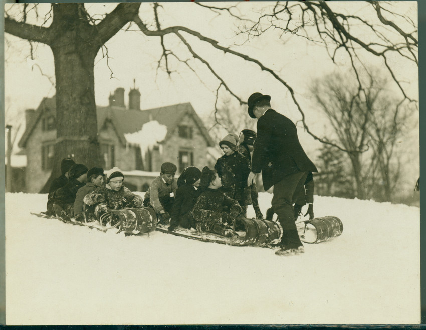 Three groups of children arrange themselves on toboggans in preparation for descending a hill. A man in a derby appears to be assisting them. There is a large house in the background.