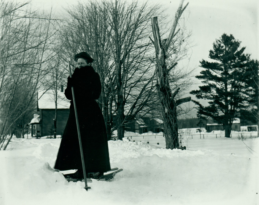 A person wearing a hat and a heavy overcoat stands on snowshoes holding a ski pole. There are houses and other buildings surrounded by woods in the background.