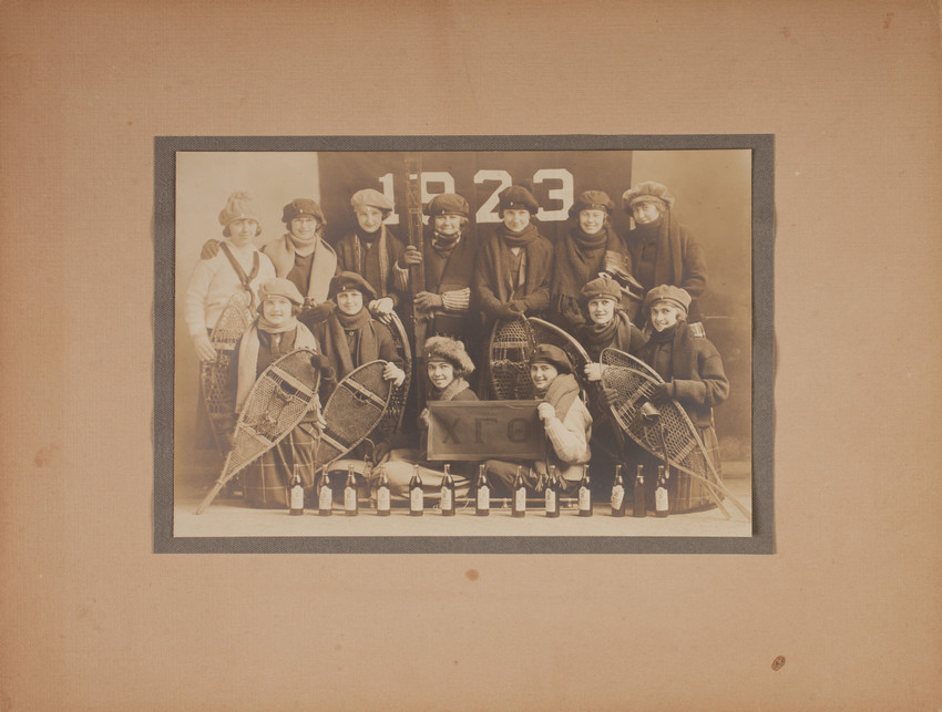 A group pf young women hold snowshoes and skiis and pose in front a flag that reads 1923.