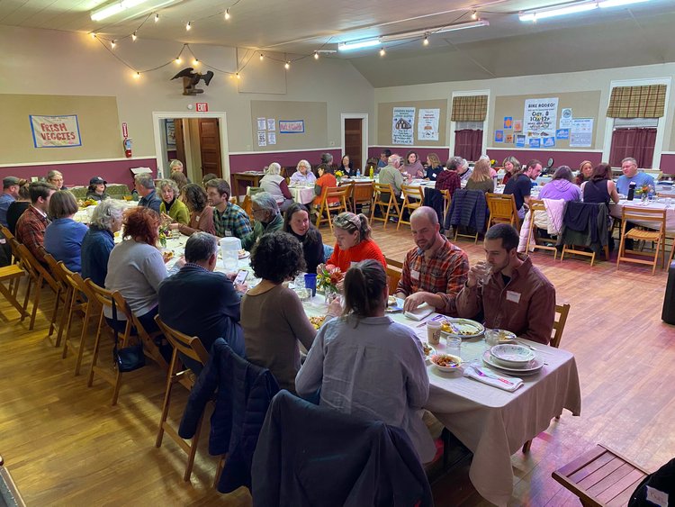 A community gathering in a room with long tables, white tablecloths, and people eating and conversing.