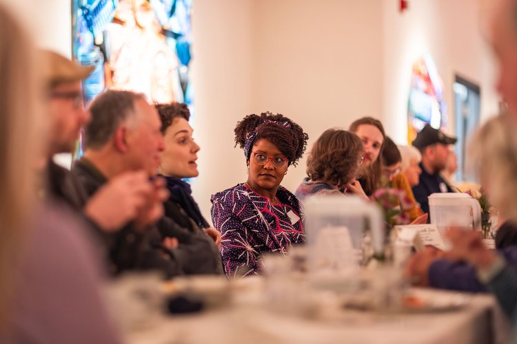 A group of people seated at a long table during an indoor event, with a woman in a patterned dress in focus.
