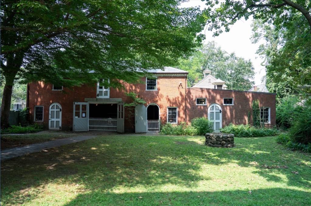 Brick building with arched and rectangular windows, surrounded by trees and lawn with a small circular stone structure in front.