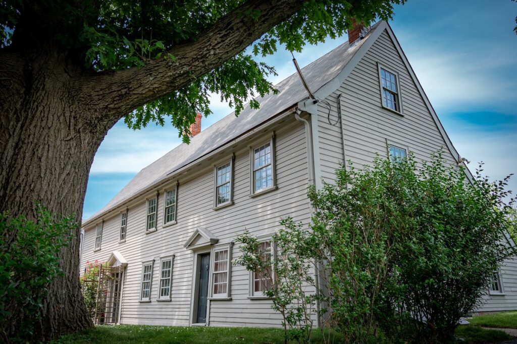 Large tree in front of an old grey wooden house with a pitched roof and multiple windows.