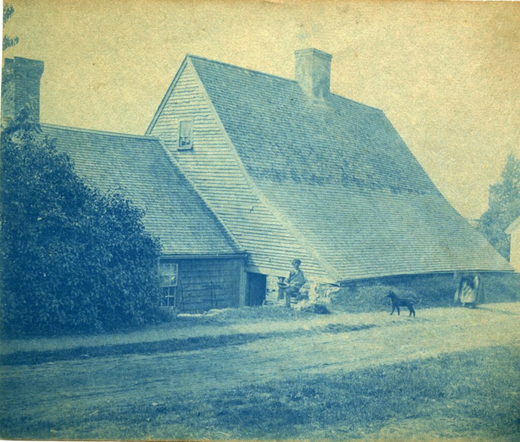 The image is a cyanotype photograph of an old farmhouse with a steeply pitched roof. In front of the house, a person is seated on the stone wall, and a dog stands on the dirt road in the foreground. Another person stands near the house, partially obscured by the building’s edge. The scene is set in a rural area with grassy surroundings.