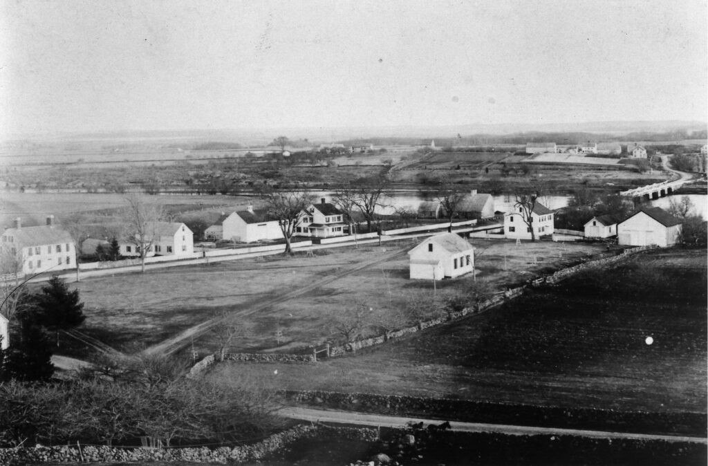 A black-and-white photograph of a rural village with houses, fields, and a bridge over a river.