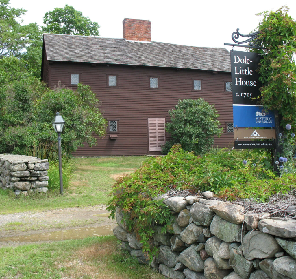 Historic brown wooden house surrounded by greenery, with a sign on the right. Transcribed Text: Dole-Little House c. 1715