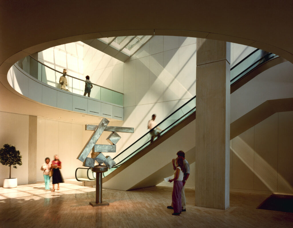 Sunlit interior with escalator, abstract sculpture, and people.