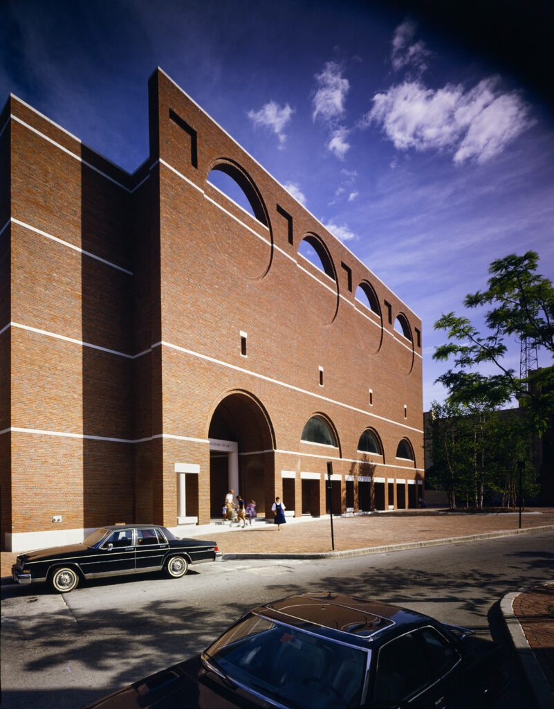 A large red brick building with arched patterns and an entrance arch, with cars parked in front. Transcribed Text: "Museum of Art"
