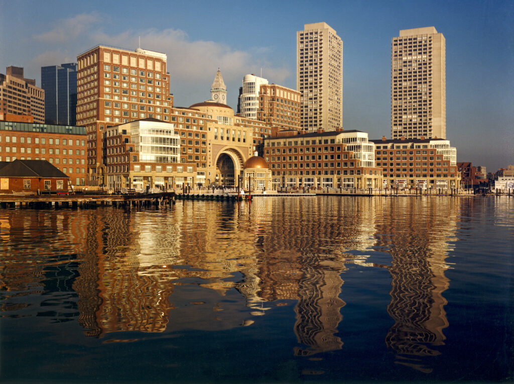 Urban skyline reflected in waterfront, featuring brick buildings and skyscrapers under a sunny sky.