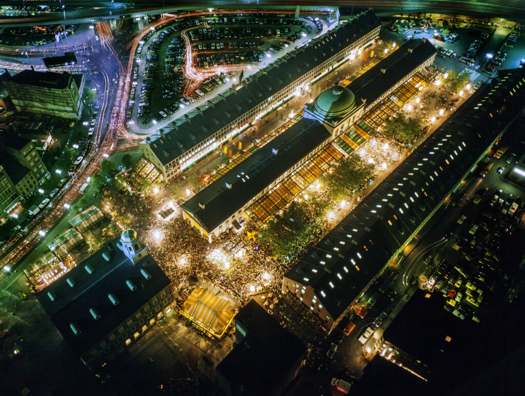 Aerial night view of a vibrant, illuminated market surrounded by parking lots and roads with light trails.
