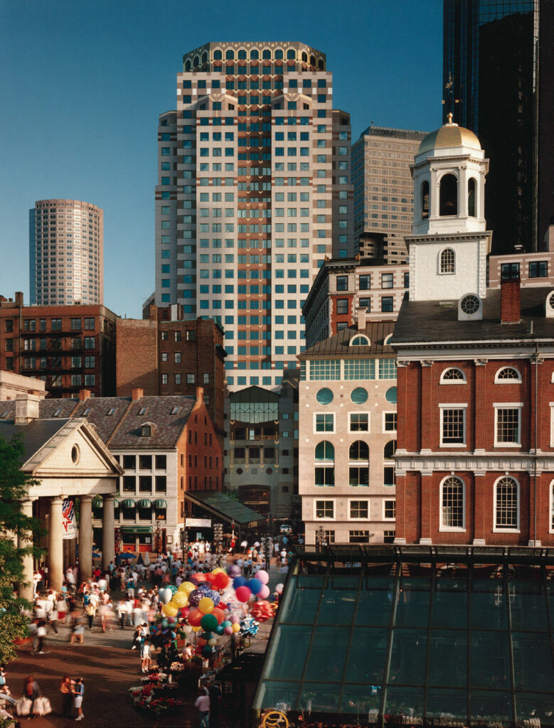 A bustling market scene with historic and modern buildings in the background, featuring balloons and a crowd of people.