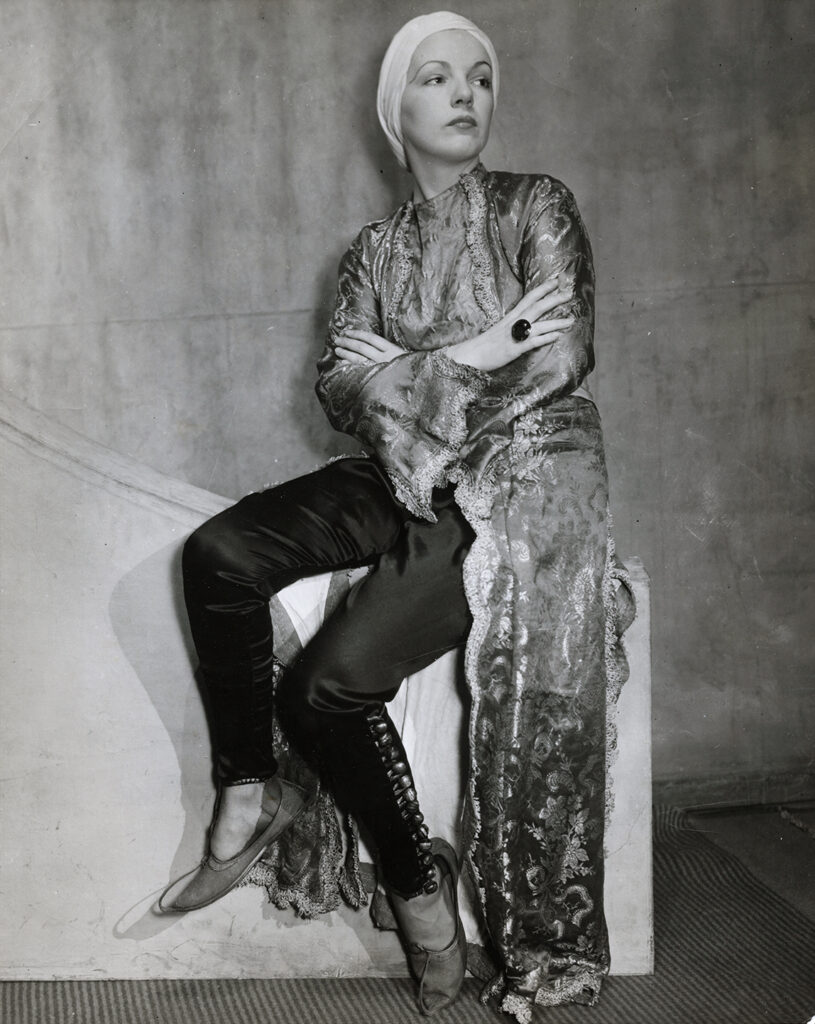 Person in an ornate robe and headscarf, sitting with arms crossed.
