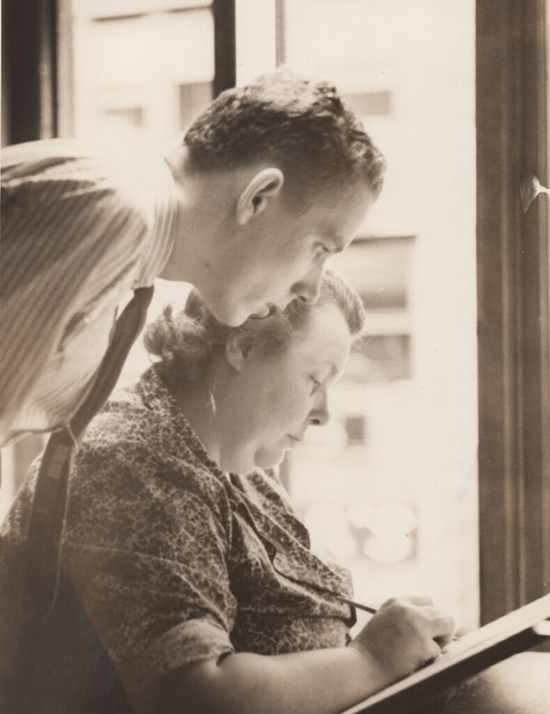 A woman writing while a man watches beside her near a window.