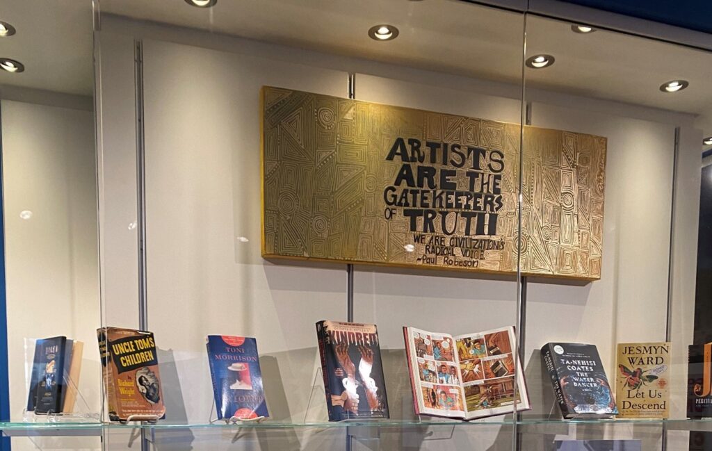 Books displayed in a well-lit glass case with a gold-toned artwork above, inscribed with a quote about artists and truth. Transcribed Text: "ARTISTS ARE THE GATEKEEPERS OF TRUTH WE ARE CIVILIZATION'S RADICAL VOICE Paul Robeson"