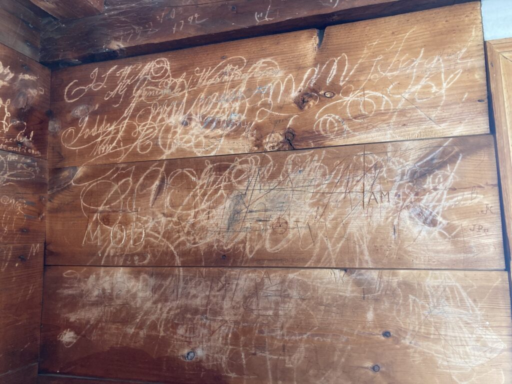 Wood wall with carved engravings and etchings in cursive and swirling patterns.
