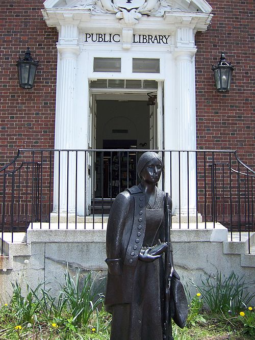 Bronze statue of a woman in front of a public library entrance with white columns. Transcribed Text: PUBLIC LIBRARY