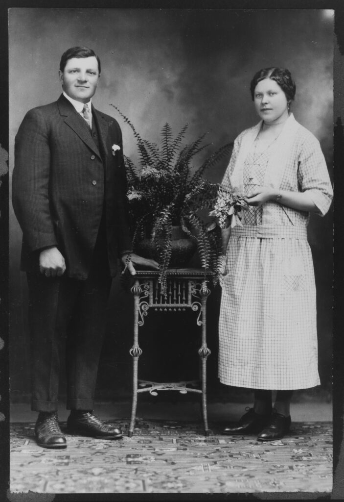 A vintage black-and-white portrait of a man in a suit and a woman in a checkered dress, standing beside a fern on a decorative table.