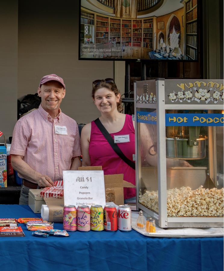 Two people smiling behind a snack table with various snacks and a popcorn machine.