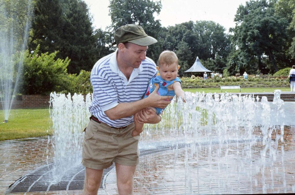 An adult holding a baby by a water fountain in a park.